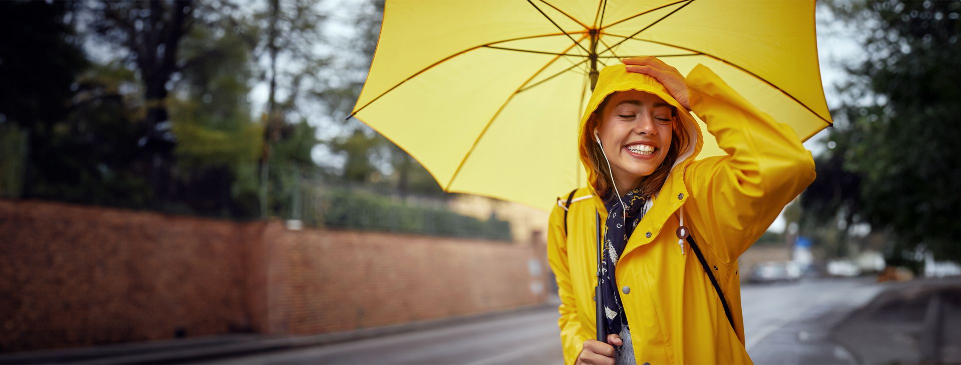 Eine lachende Frau steht am Gehsteig im Regen und hält einen aufgespannten Regenschirm in der Hand.